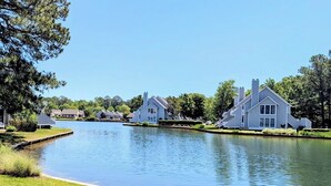 A lakeside view with modern houses, a calm water body, and two Adirondack chairs. - Ground-Floor Sea Colony Condo. Pond Views. Resort Amenities! (Bethany Beach)