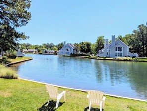 A lakeside view with modern houses, a calm water body, and two Adirondack chairs. - Ground-Floor Sea Colony Condo. Pond Views. Resort Amenities! (Bethany Beach)