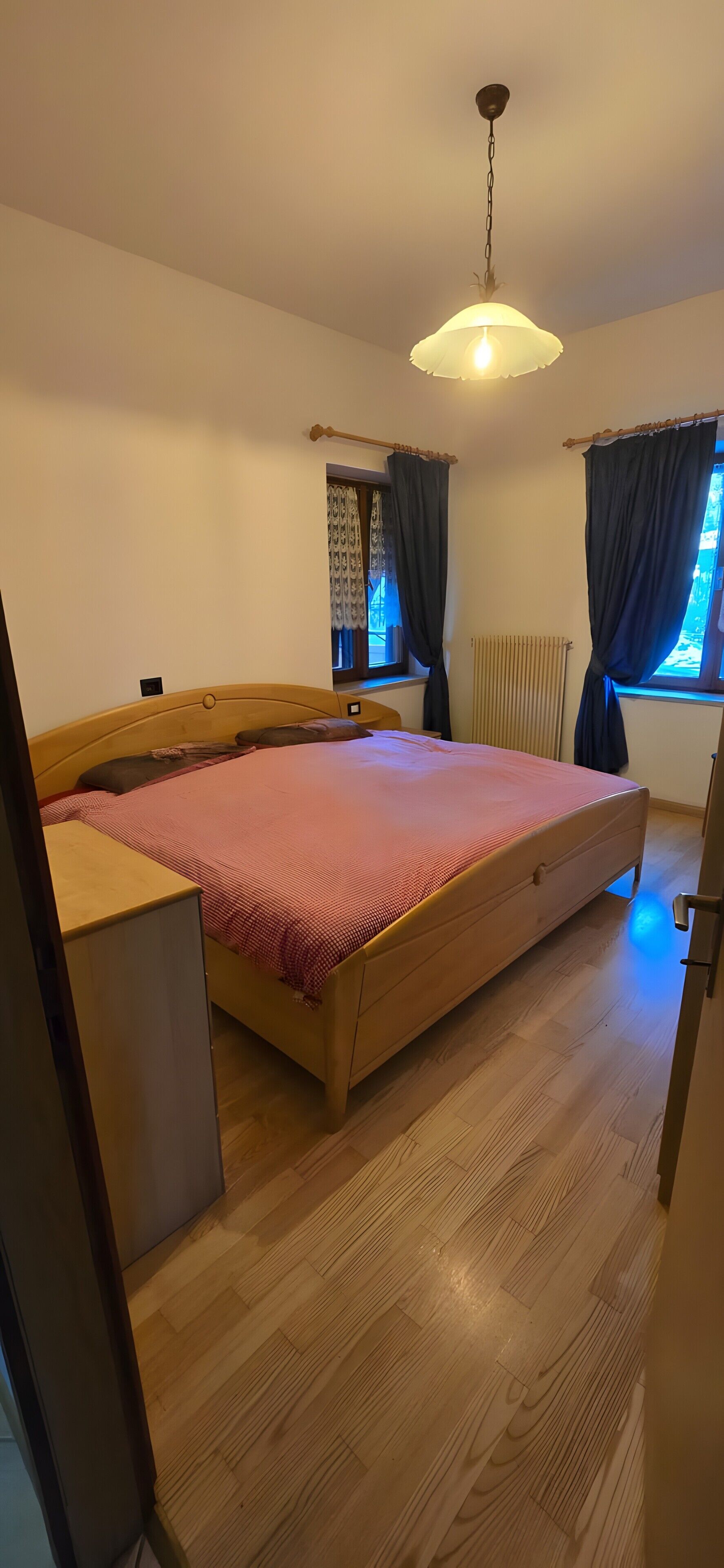 A bedroom with a wooden bed, a hanging light fixture, a window with curtains, and a radiator.