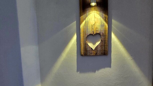Un rincón de una habitación con una lámpara fijada a la pared que proyecta sombras, un pequeño banco con cojín y un suelo de baldosas.
