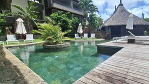 A clear pool with a wooden deck, lounge chairs, and thatched-roof structures. - Luxury Furnished Apartment in Grand Baie (Grand Baie)