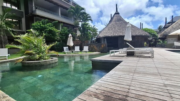 A resort pool area with a wooden deck, lounge chairs, and thatched-roof huts. - Luxury Furnished Apartment in Grand Baie (Grand Baie)