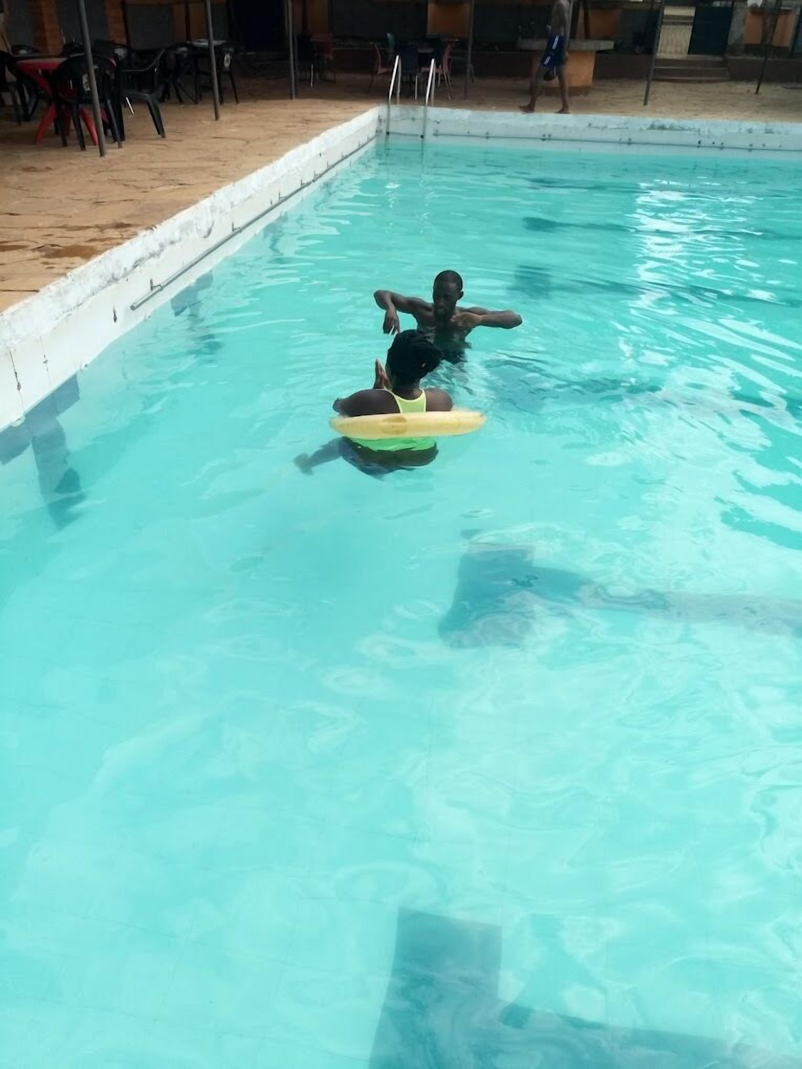A swimming pool with clear blue water, a person floating on a yellow float, and a few chairs and tables around the pool area.