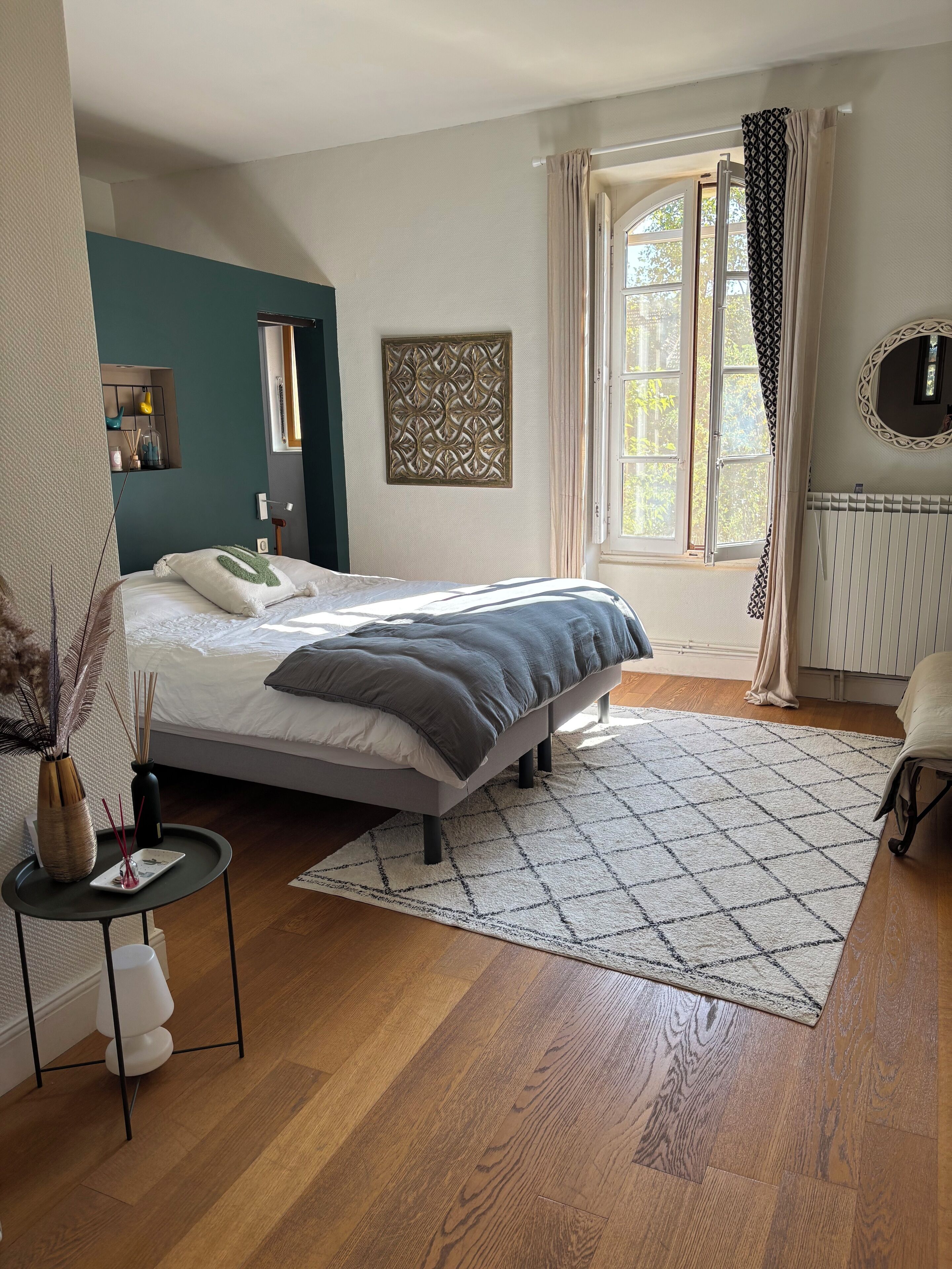 Une chambre à coucher comprenant un lit, une petite table de chevet, un élément décoratif mural, une fenêtre avec des rideaux et un radiateur.
