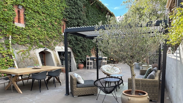 Um pátio externo com mesa e cadeiras de madeira, mobiliário de vime e plantas em vasos.