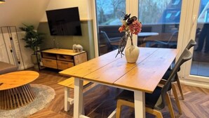 Zona pranzo con tavolo e sedie in legno, un vaso con fiori e un televisore a parete.