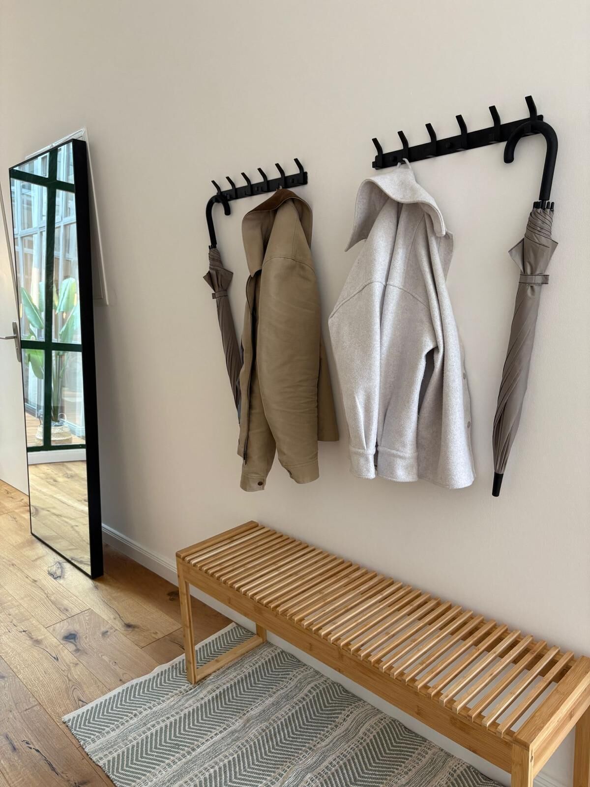 A room with a wooden bench, a coat rack with two coats and two umbrellas, and a large mirror on the wall.