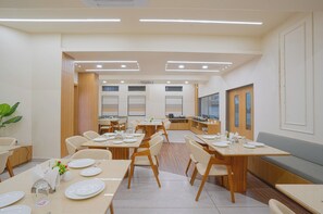 Uma sala de jantar moderna com várias mesas e cadeiras, uma cozinha ao fundo e uma janela grande que oferece luz natural.