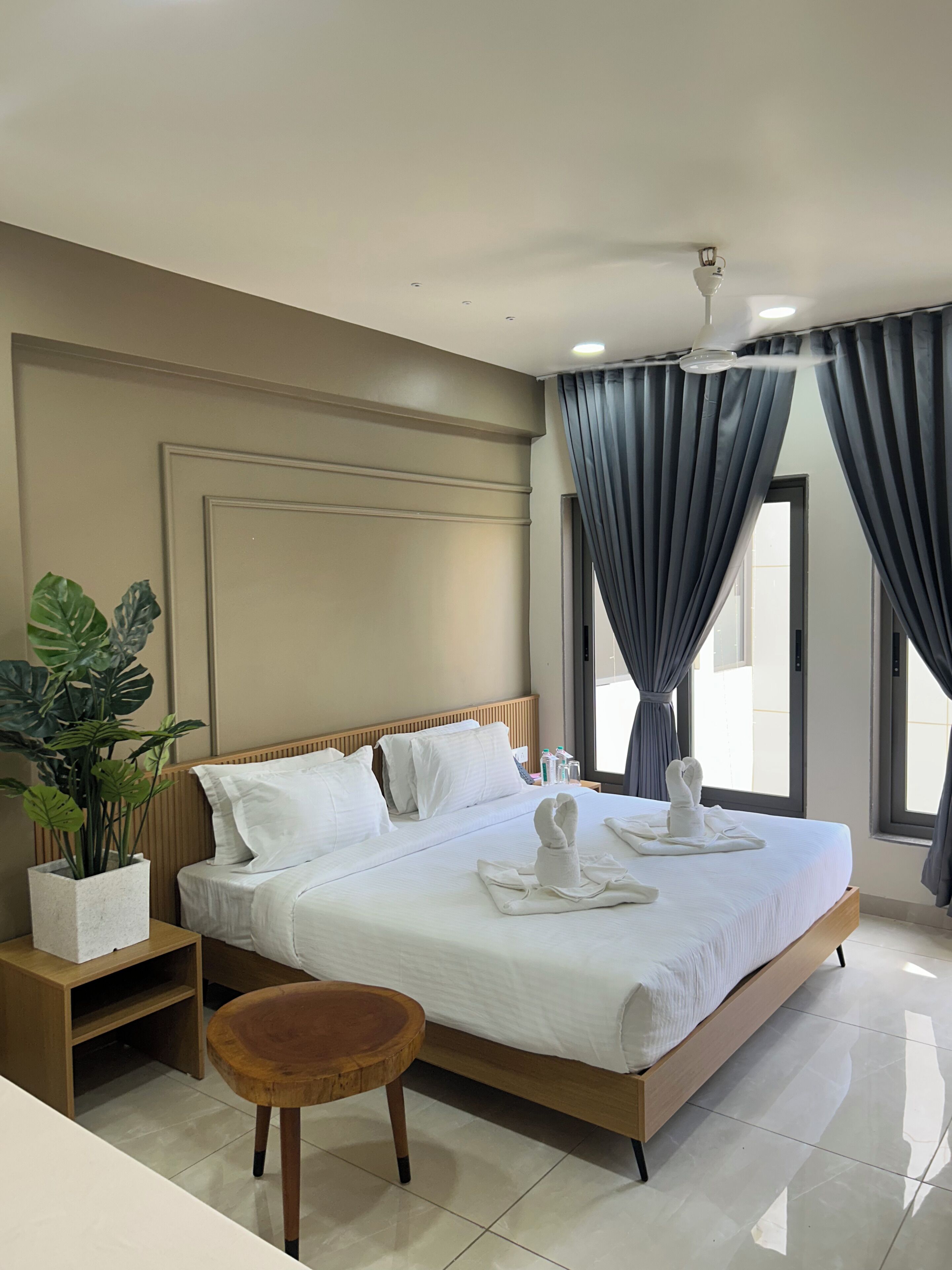 Une chambre d’hôtel avec un grand lit, une table de chevet, un petit tabouret en bois, une plante en pot et une vue sur l’extérieur par la fenêtre.