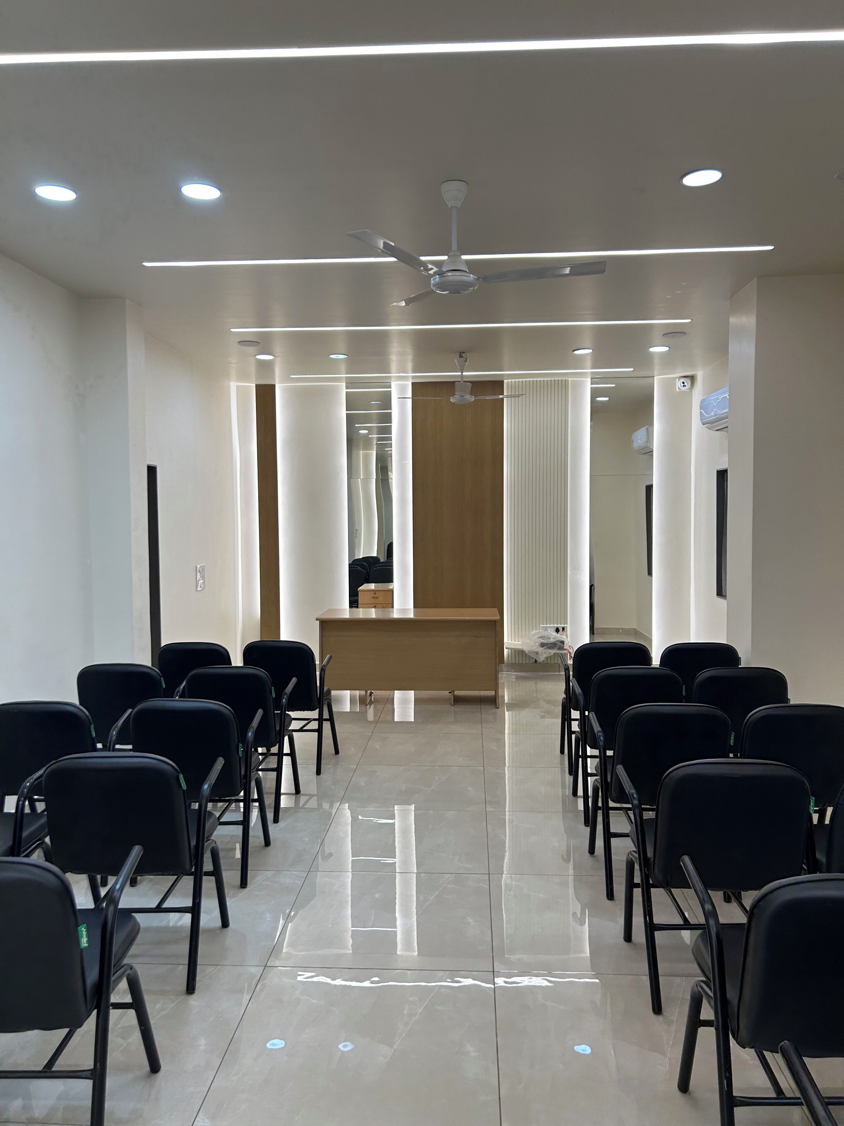 Un salón de conferencias con mostrador de recepción, sillas negras dispuestas en filas, ventilador de techo e iluminación empotrada.