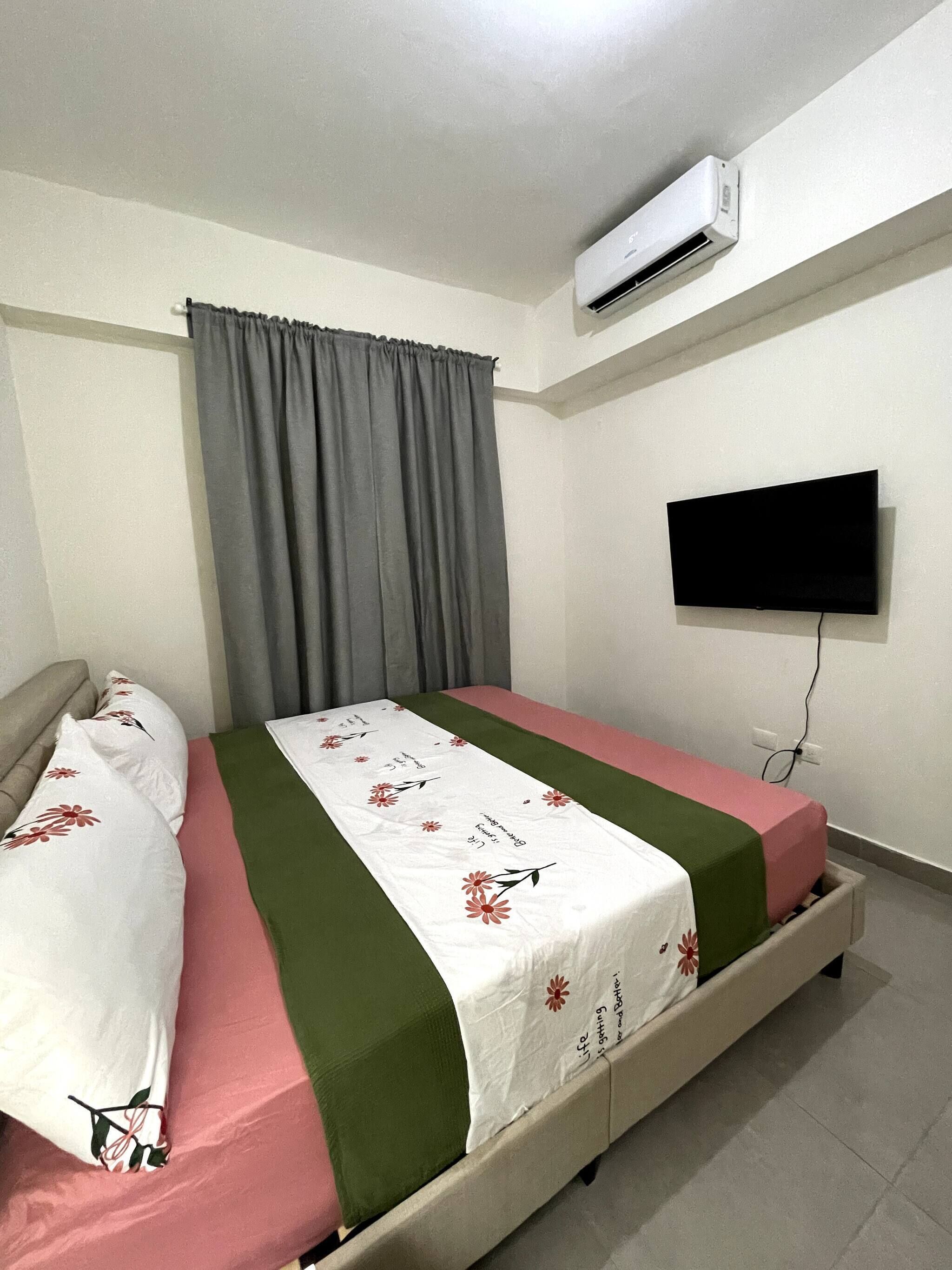 A single bed with a floral bedspread, a grey curtain, an air conditioning unit, and a TV mounted on the wall.