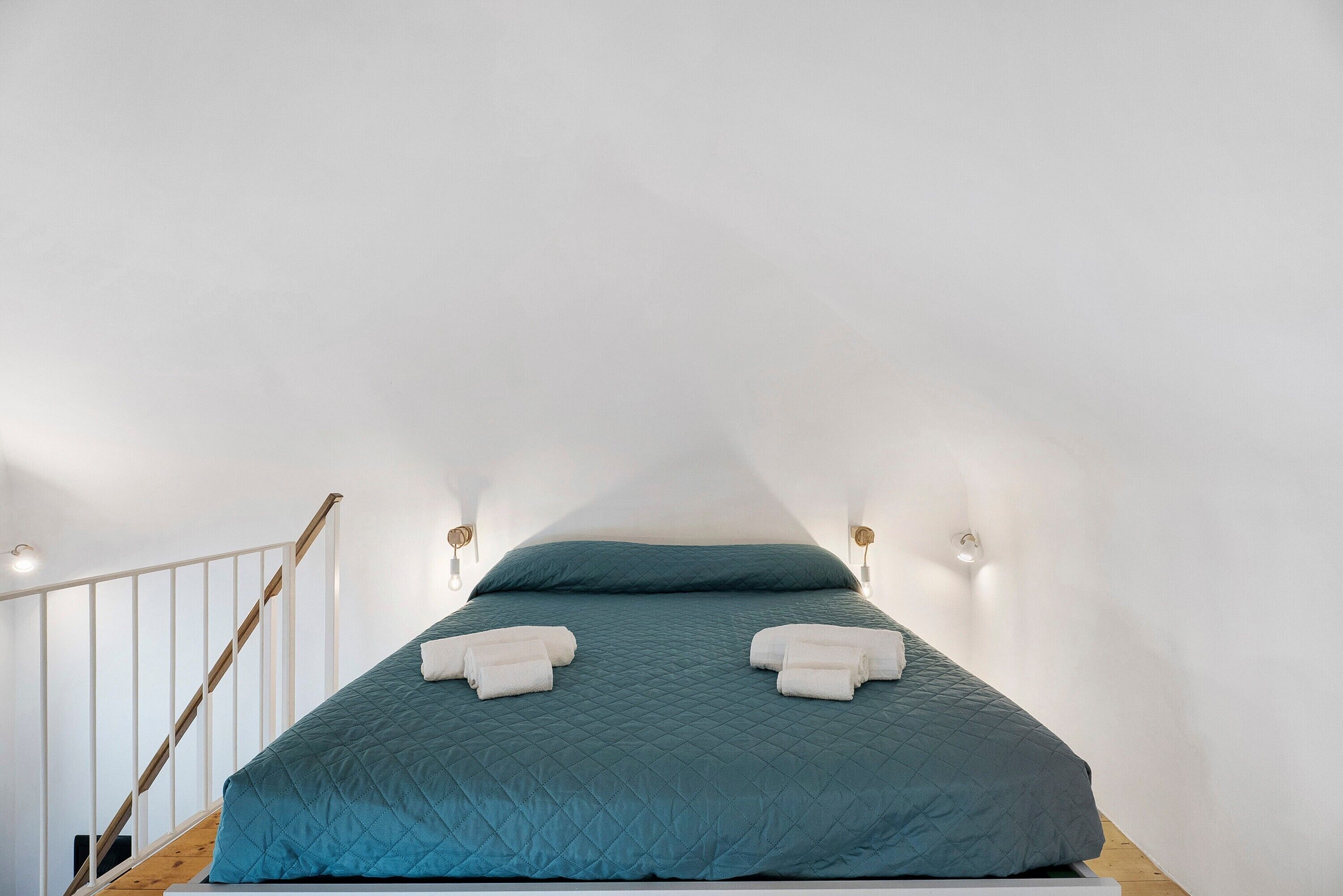 A single bed with a teal quilt, two white towels, a wooden staircase, and wall-mounted sconces.