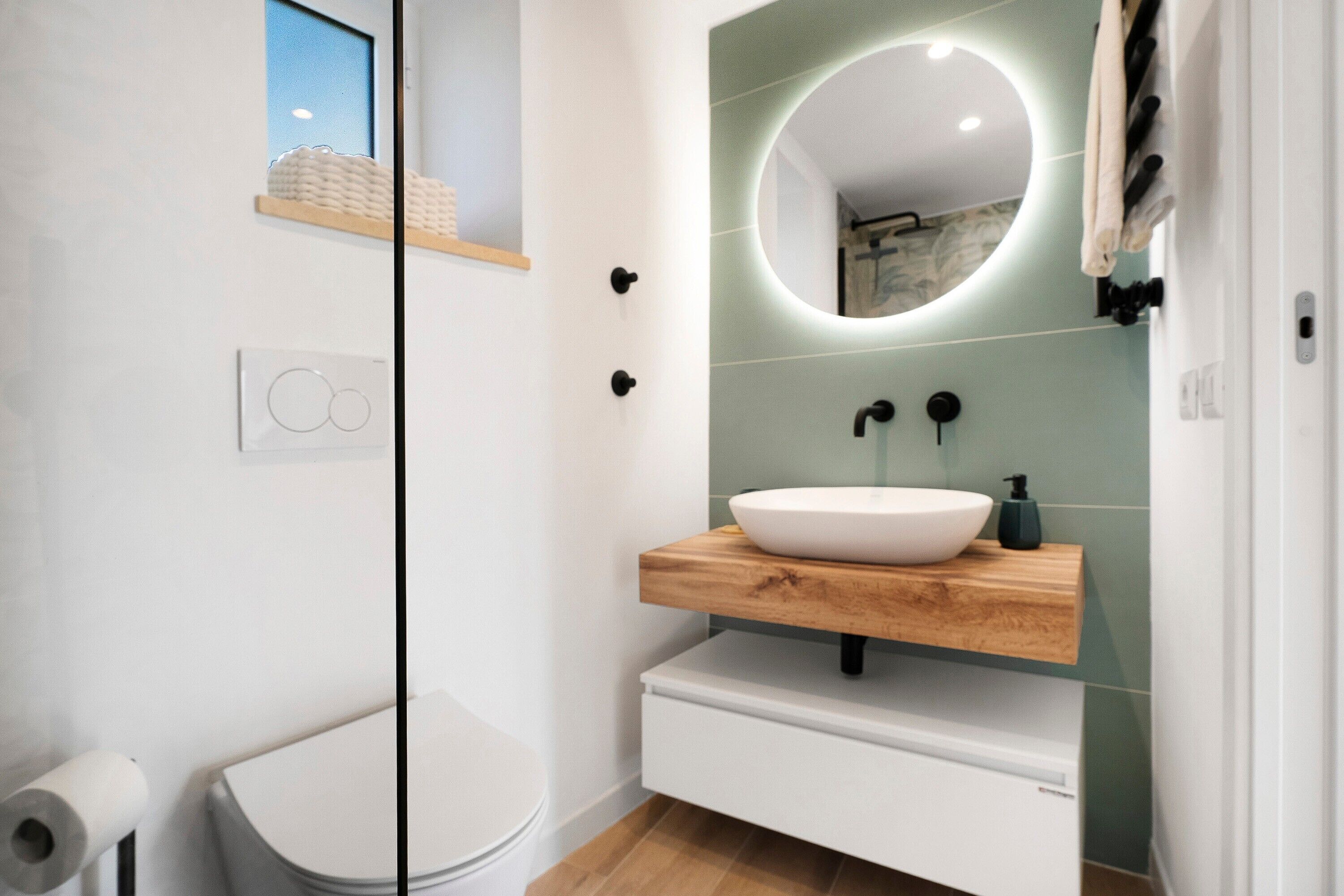 A modern bathroom with a white toilet, a wooden vanity with a white sink, a round mirror with a light, and a towel rack.