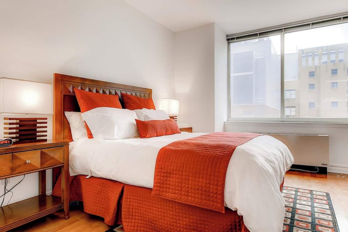 Habitación de hotel con una cama grande, una mesita de noche con lámpara, una ventana con vistas a edificios y suelo de madera.