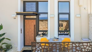 Une façade moderne avec une porte en bois, deux fenêtres donnant sur l’océan et un petit espace extérieur aménagé avec des chaises jaunes.