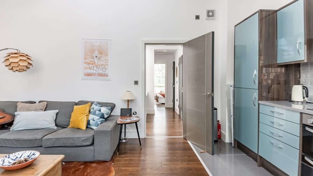 A modern living room with a grey sofa, a small table with a bowl, and a lamp. A hallway leads to a kitchen with blue cabinets and a stove. - Two bedroom loft in central Bristol (City of Bristol)