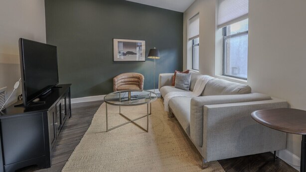 Una sala de estar moderna con una pared de acento verde oscuro, un sofá blanco, una mesa de centro de vidrio, un sillón de cuero, un televisor sobre un soporte negro y una mesita redonda.