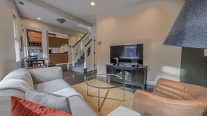 Una sala de estar moderna con un sofá, un sillón y una mesa de centro de vidrio. Un televisor de pantalla plana está montado en un mueble oscuro, y se ve una escalera al fondo que conduce a un piso superior.