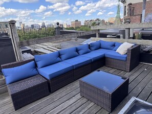 Terraza en la azotea con un amplio sofá modular, un pequeño reposapiés, suelo de madera y vistas a un horizonte urbano.
