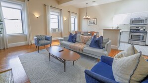 Ein Wohnzimmer mit Sofa, Sessel und Couchtisch, eine Küche mit Geräten und große Fenster mit Vorhängen.
