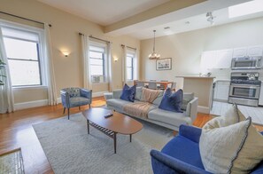 Un salon avec un canapé, un fauteuil et une table basse, une cuisine équipée et de grandes fenêtres avec des rideaux.