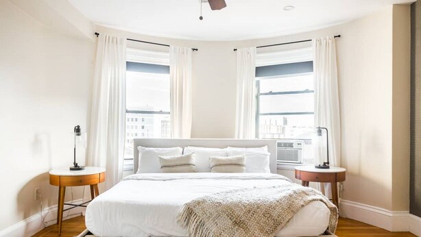 A modern bedroom with a large bed, two bedside tables, and a view of the city through large windows. - The only 4 Bedroom Apartment w/ Roofdeck in Boston!  8min from Fenway Park, close to universities! (Boston)