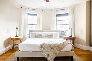 Un dormitorio moderno con una cama grande, dos mesitas de noche y vistas a la ciudad a través de amplias ventanas.