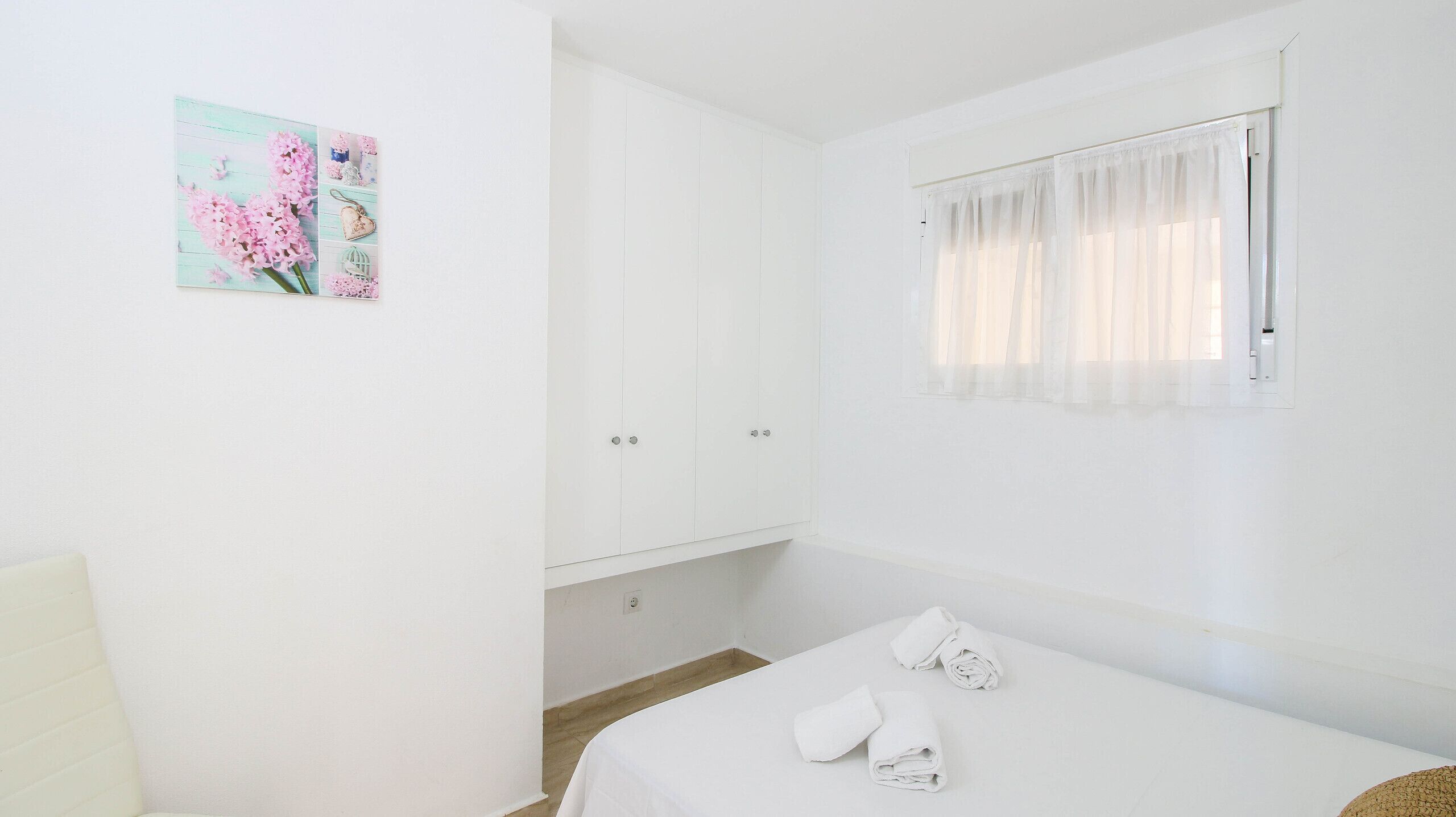 Une chambre à coucher blanche, avec un lit, une fenêtre ornée de rideaux légers et un meuble fixé au mur.