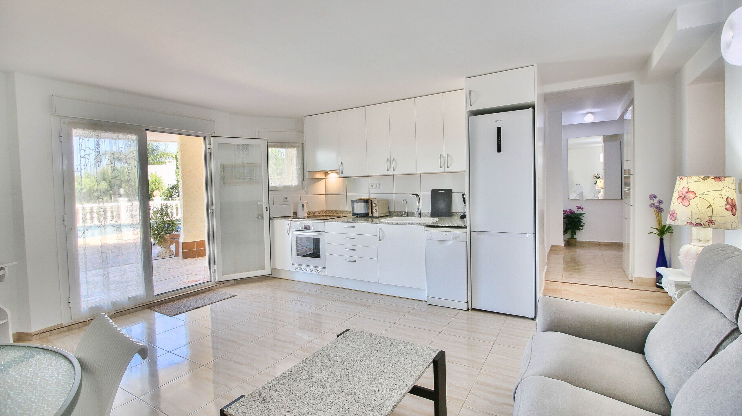 A modern kitchen with white cabinets, a built-in oven, and a refrigerator. There is a dining area with a table and chairs, and a sofa in the living room.