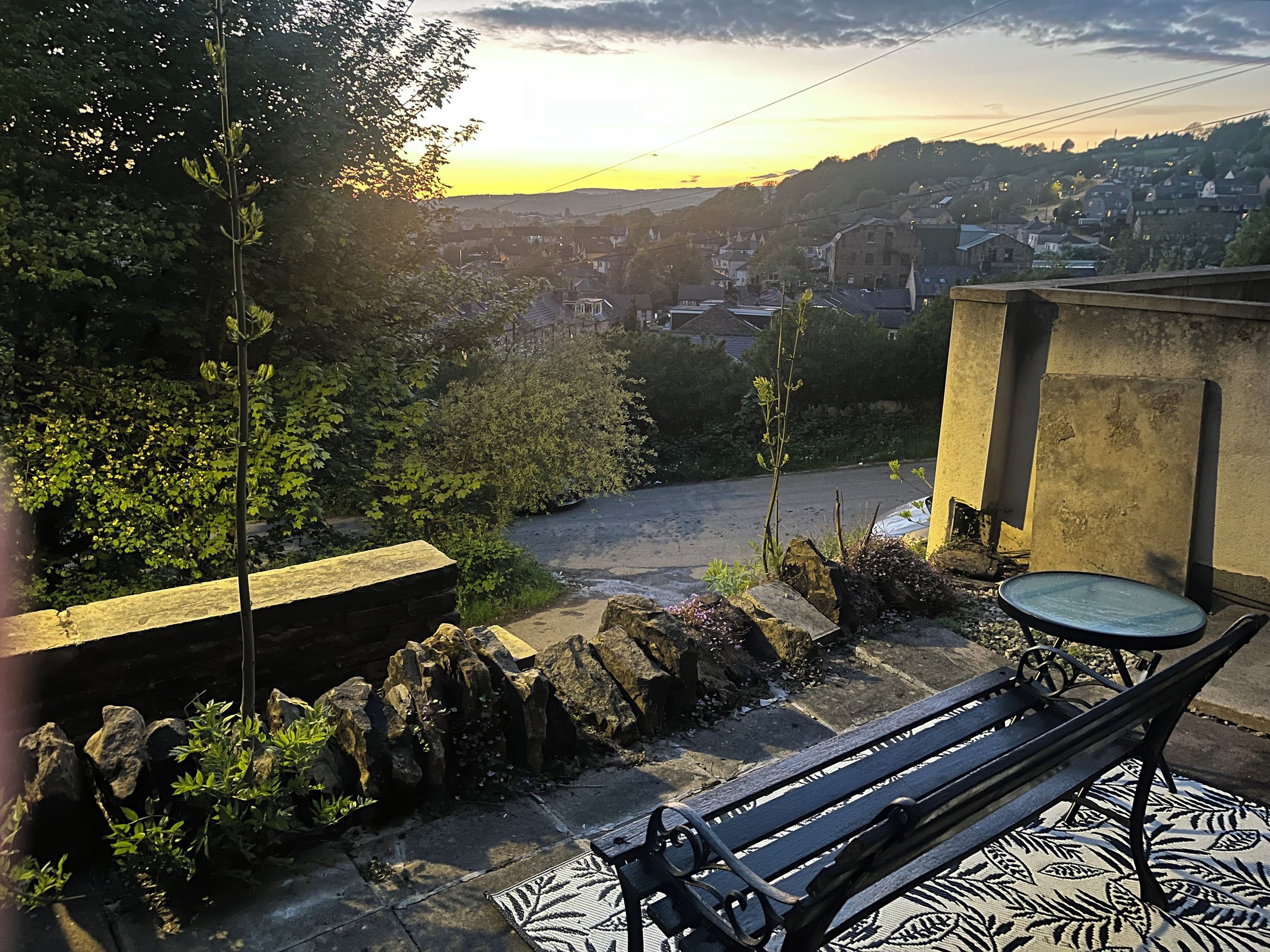 Eine Bank mit gemustertem Unterleger, eine Steinmauer und Blick auf ein Wohngebiet bei Sonnenuntergang.