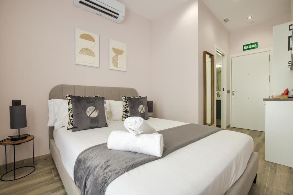 Una cama bien hecha con una manta gris y una toalla blanca enrollada, una lámpara de noche y dos obras de arte colgadas en la pared.