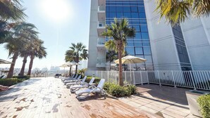 A modern building with a reflective glass facade, a rooftop pool area with sun loungers, and palm trees. - StayShort - Studio in Damac Hills that Sleeps 3 with Terrace! (Damac Hills)