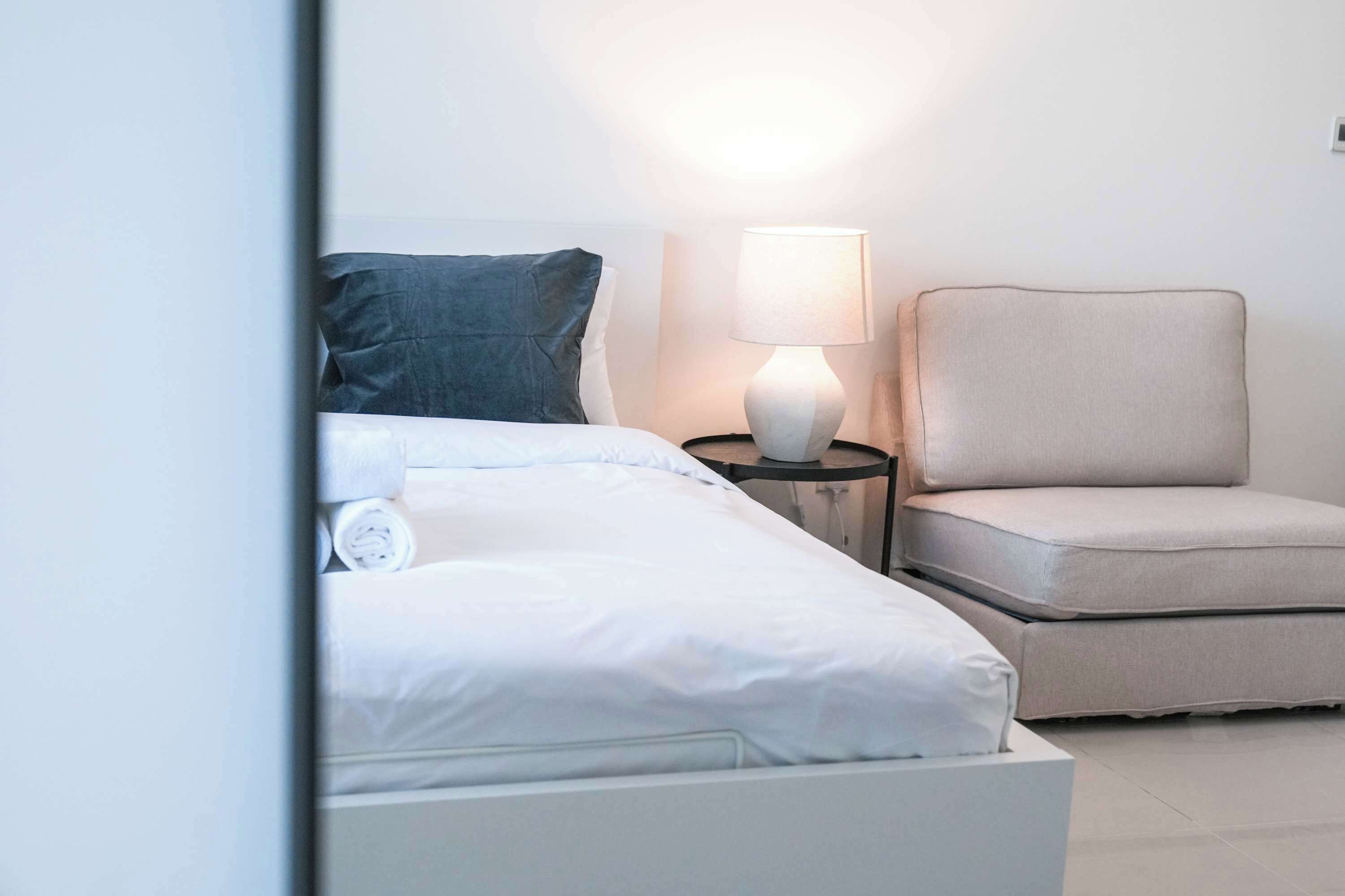 A neatly made bed with white linens and a dark blue pillow, a bedside table with a lamp, and a light-colored armchair.
