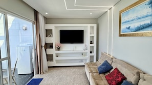 A modern living room with a flat-screen TV, a beige sofa with patterned cushions, and a white entertainment unit with shelves. - Duplex penthouse in the Porto Real Condominium.   (Mangaratiba)