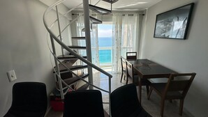 Une pièce dotée d’un escalier en colimaçon menant à un balcon offrant une vue sur la mer. On y trouve une table à manger avec des chaises, ainsi qu’une petite table sur laquelle est posé un ordinateur portable.