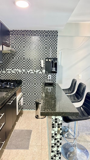 A modern kitchen with a black and white tiled backsplash, a stainless steel stove, and a bar area with a glass top and black chairs. - Duplex penthouse in the Porto Real Condominium.   (Mangaratiba)