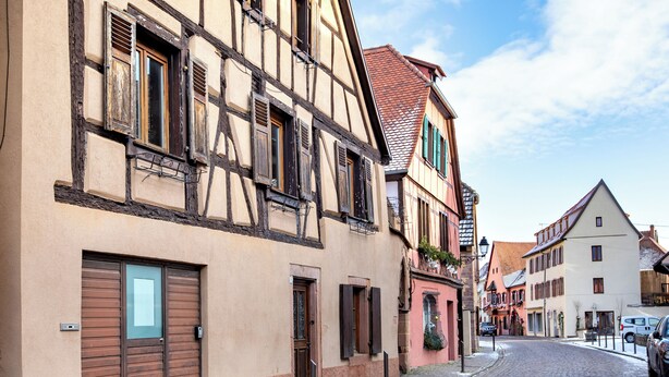 Une rue pavée bordée de bâtiments traditionnels à colombages, avec des volets en bois et un mélange de portes ouvertes et fermées.