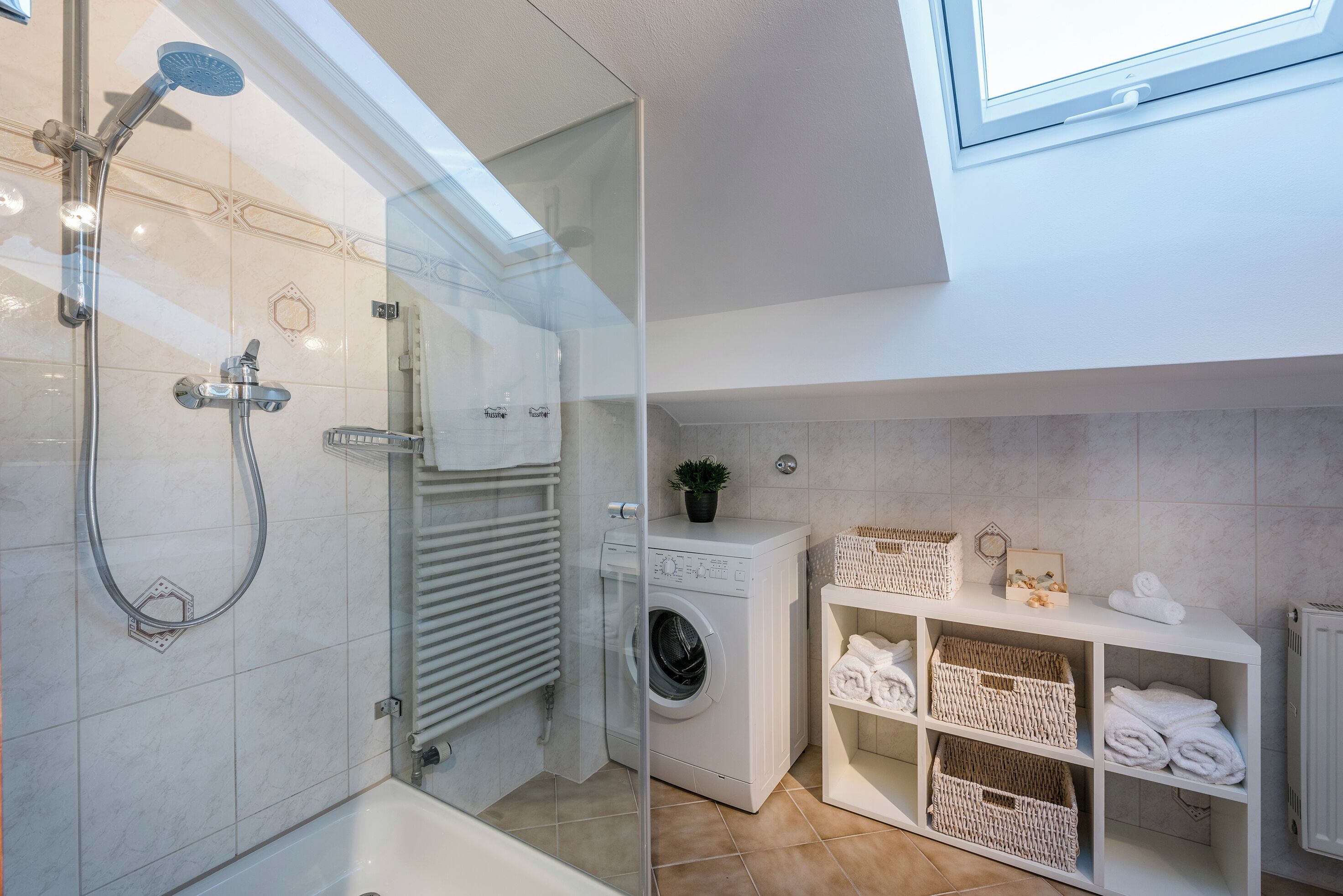 A modern bathroom with a glass-enclosed shower, a built-in washing machine, and a shelf with neatly folded towels.