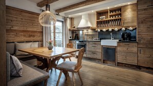 A modern kitchen with wooden beams, a dining area with a wooden table and chairs, and built-in shelves with kitchenware. - Hussnhof Farm Lodge (Sachsenkam)