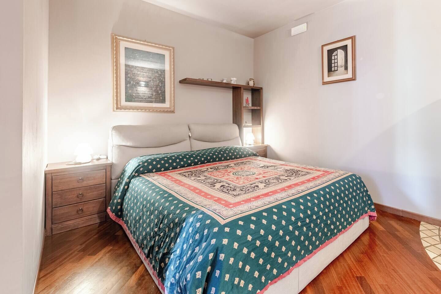 A bedroom with a bed, a nightstand, a shelf with decorative items, and framed pictures on the wall.