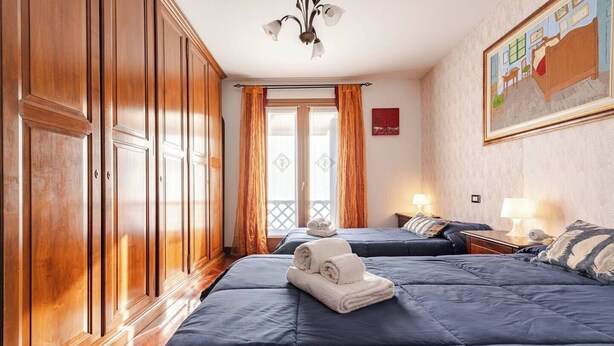 Habitación de hotel con dos camas, armarios de madera, un balcón con cortinas naranjas y un cuadro en la pared.