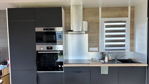 A compact kitchen with a built-in oven, a microwave, and a stovetop. There is a window with blinds, a sink, and a small countertop with a bottle and a cup. - Charming chalet SAINT JACQUES SARZEAU GOLFE DU MORBIHAN - 330m² private garden (Sarzeau)