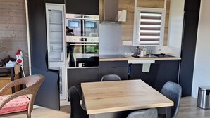 A compact kitchen with a built-in oven, a sink, and a window with blinds. There is a dining area with a table and chairs. - Charming chalet SAINT JACQUES SARZEAU GOLFE DU MORBIHAN - 330m² private garden (Sarzeau)