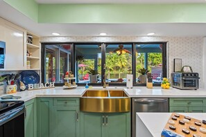 Eine Küche mit grünen Schränken, einer Edelstahl-Spüle, einem eingebauten Backofen und einem großen Fenster mit Blick auf eine Außenbestuhlung.