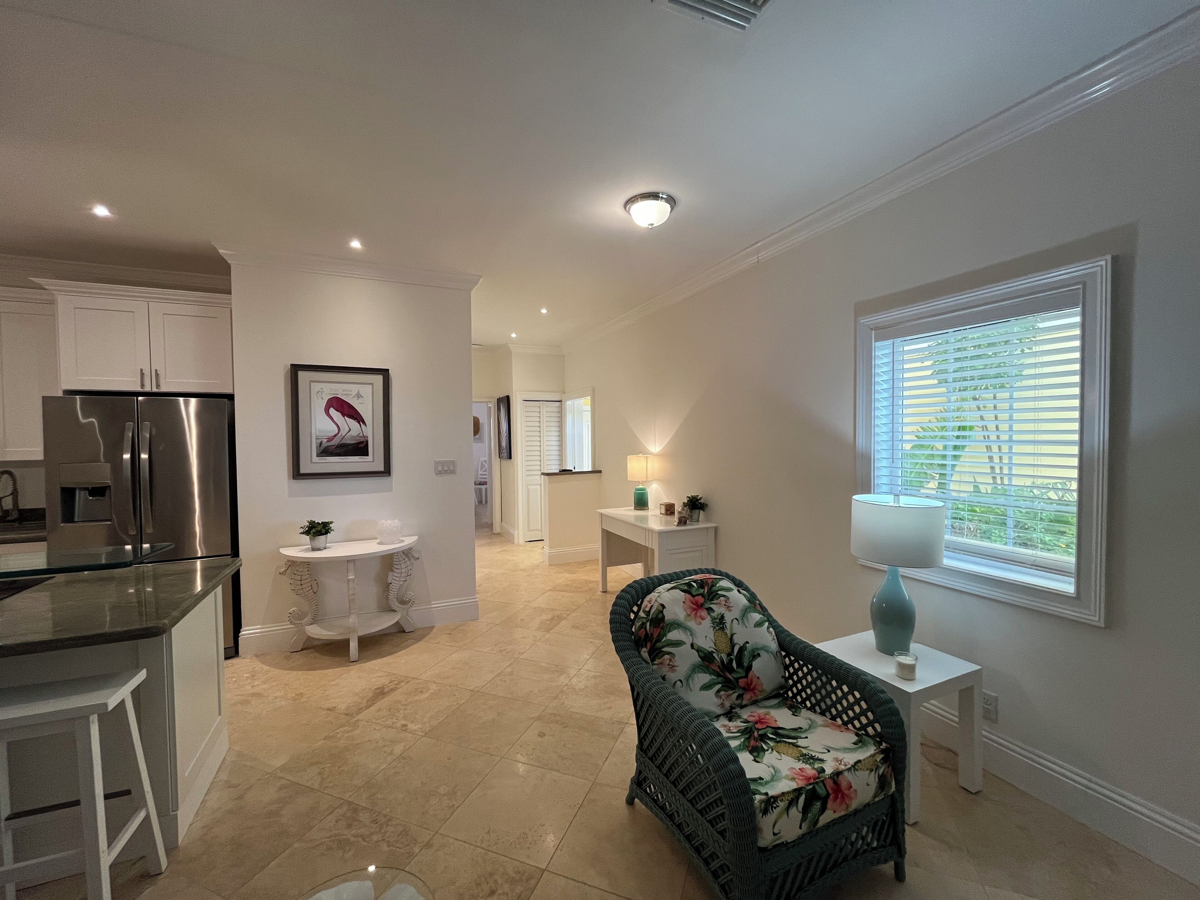 A modern kitchen with a stainless steel refrigerator, a dining area with a glass table, and a living space with a wicker chair and a window with blinds.