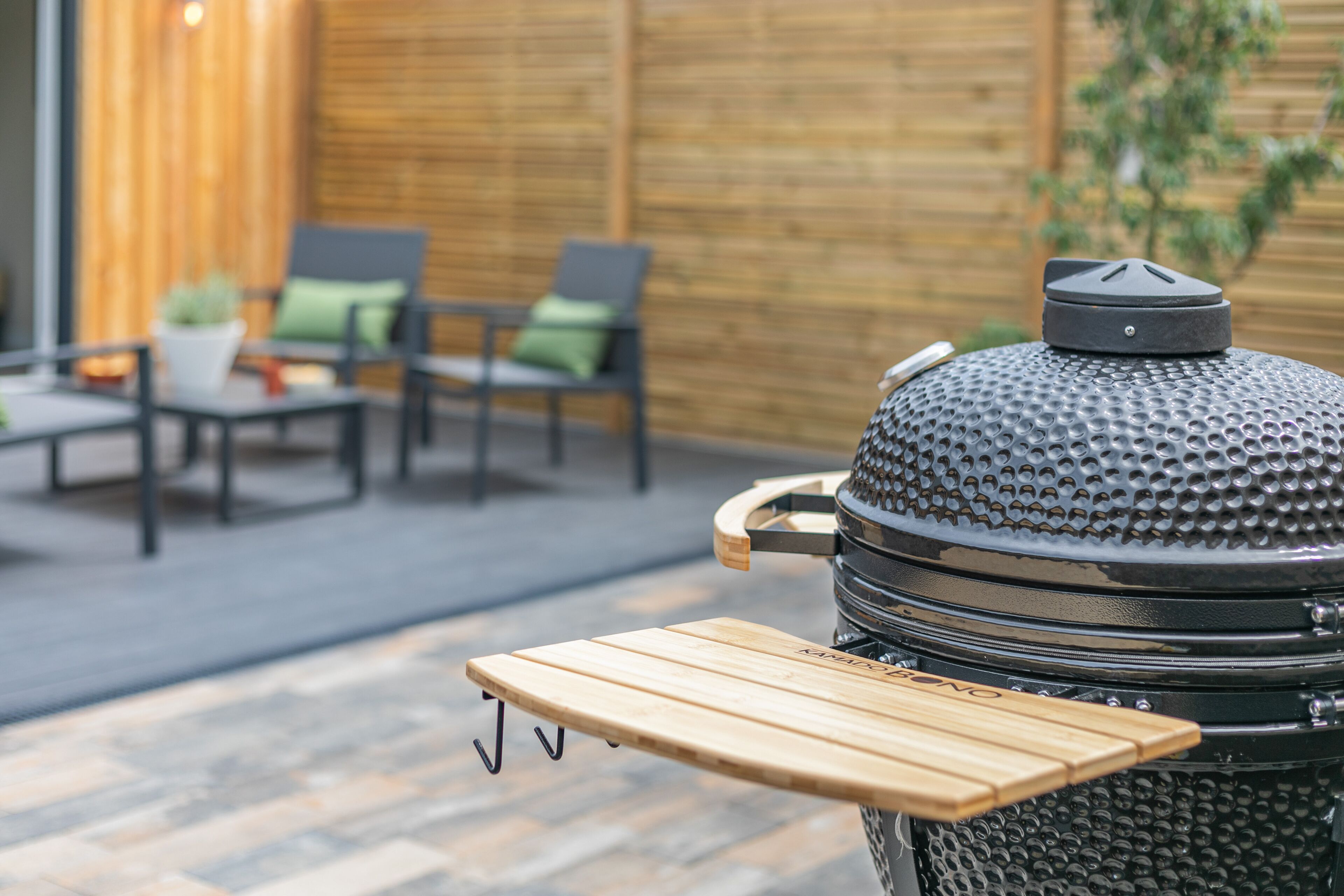 Una parrilla para barbacoa con una bandeja de madera y una tapa de metal, ubicada en un patio con sillas y un muro de madera al fondo.