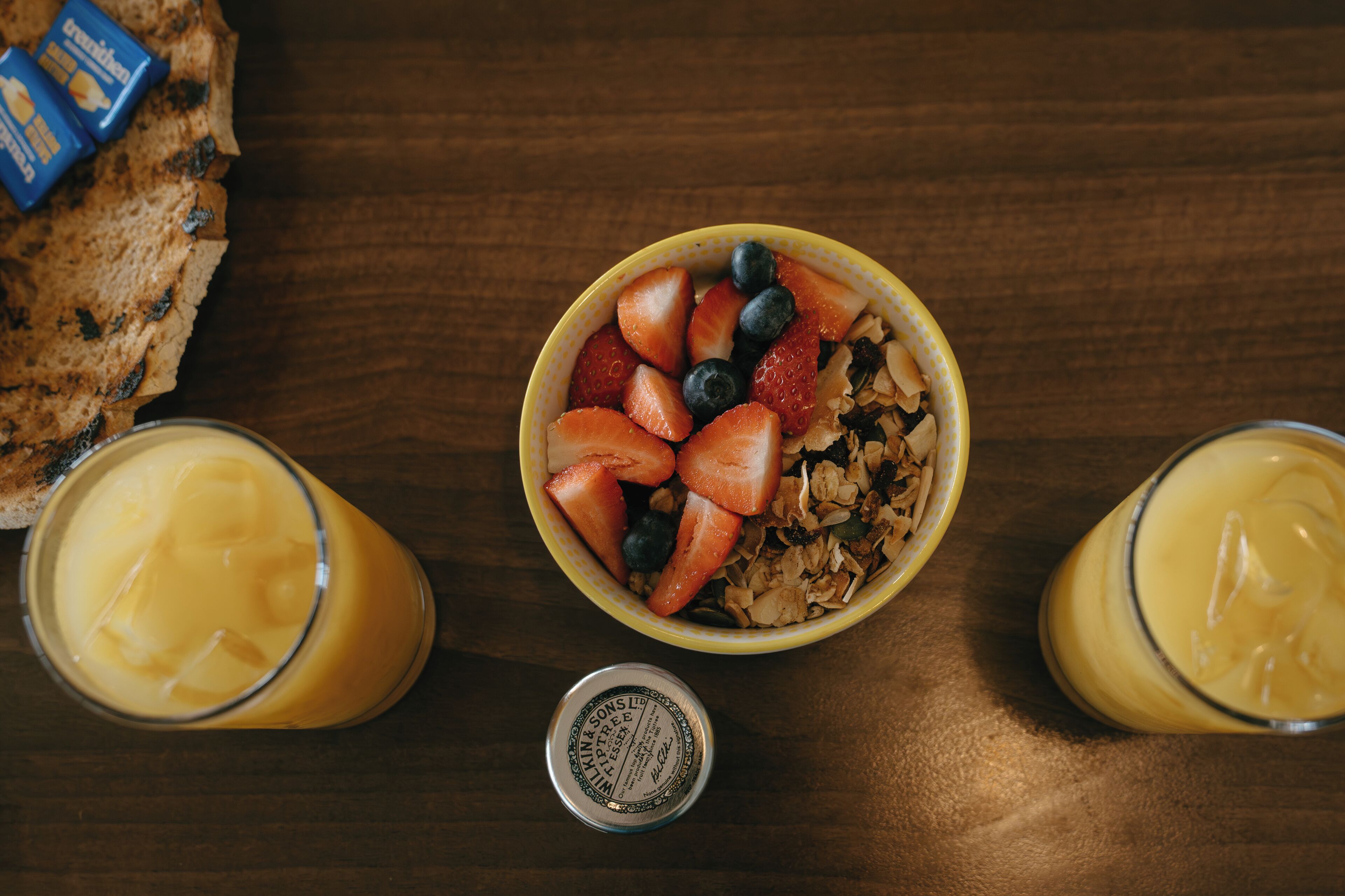 Desayuno con un tazón de cereal, fresas, arándanos y granola, acompañado de dos vasos de jugo de naranja y un frasco pequeño de miel.
