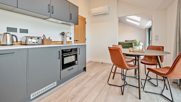 A modern kitchen with grey cabinets, a built-in oven, and a dining area with orange chairs and a round table. - Landal Whalesborough (Bude)
