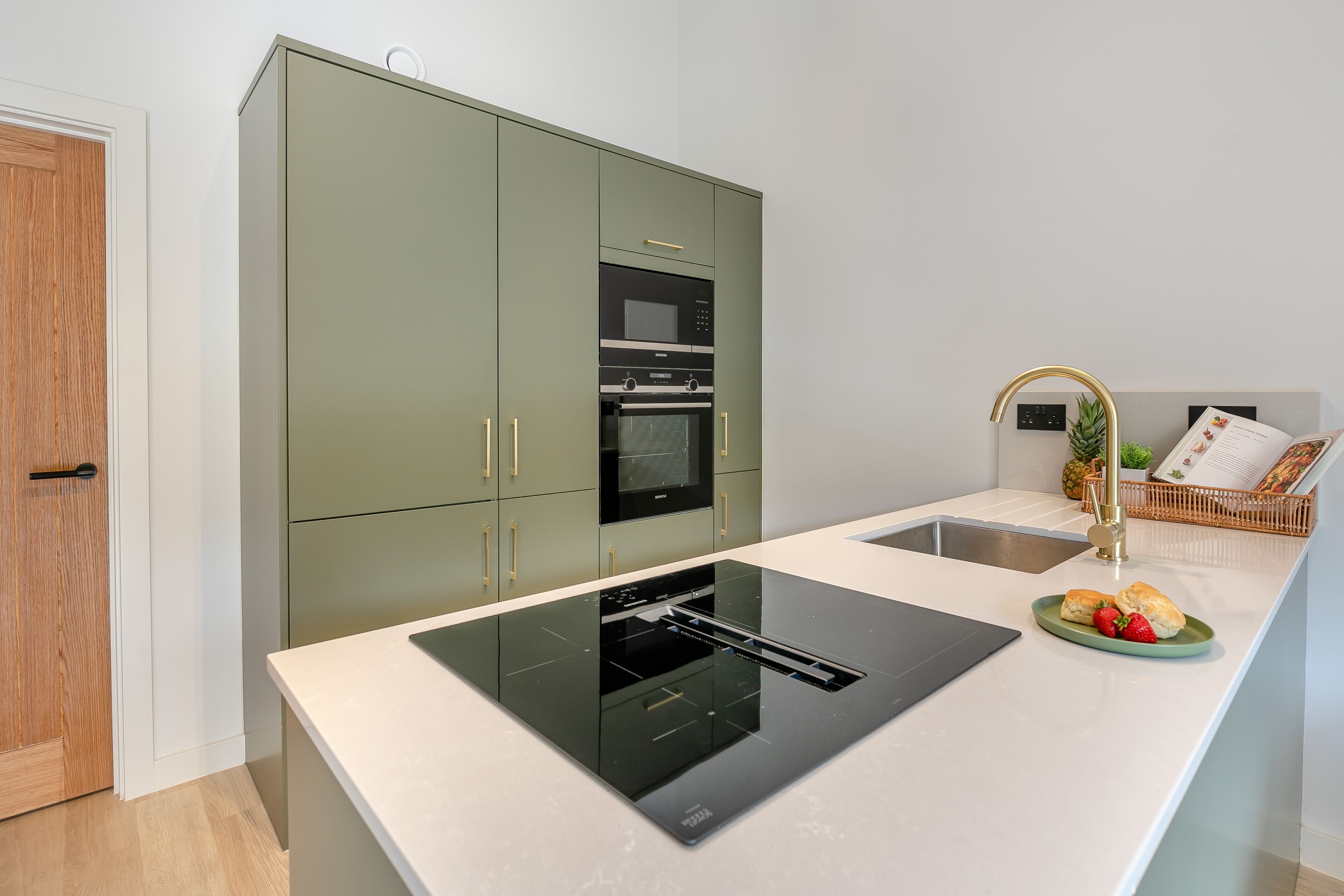 A modern kitchen with a built-in oven and microwave, a large island with a sink, and a small dining area with a basket of bread and fruit.