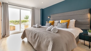 A modern bedroom with a large bed, a view to the outdoors, and a small side table with a plant. - Landal Whalesborough (Bude)